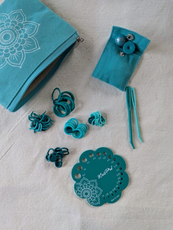 Photo d’accessoires de tricot KnitPro Mindful posés sur un tissu beige : marqueurs, jauge d’aiguilles, aiguilles à repriser et petite pochette turquoise assortie. Accessoires inclus dans le kit d’aiguilles circulaires interchangeables KnitPro Mindful Believe.