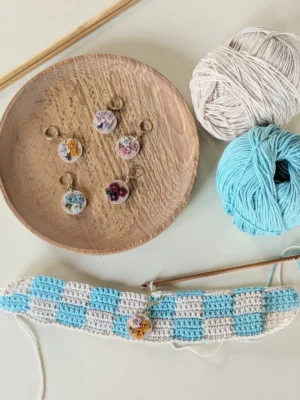 photo d'un ouvrage en crochet avec un marqueur de mailles en bois décoré de fleurs en micro crochet, à l'arrière plan une assiette en bois dans lequel se trouve d'autres marqueurs de mailles en bois et des pelotes de laine