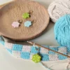 marqueur de maille sur un ouvrage en crochet beige et bleu. A l'arrière plan deux pelotes de coton et un support en bois sur lequel est posé 3 autres marqueurs de mailles en forme de fleur.