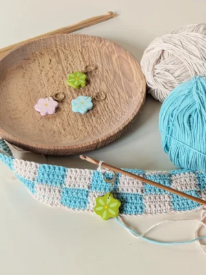 marqueur de maille sur un ouvrage en crochet beige et bleu. A l'arrière plan deux pelotes de coton et un support en bois sur lequel est posé 3 autres marqueurs de mailles en forme de fleur.