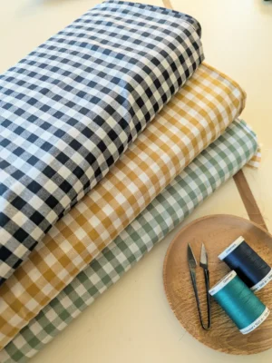 tissu vichy enroulé de couleur bleu marine, jaune et vert menthe en premier plan il y a deux bobines de fils de couture et ciseaix coupe-fil