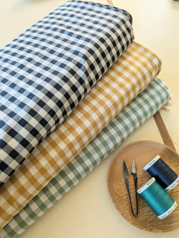 tissu vichy enroulé de couleur bleu marine, jaune et vert menthe en premier plan il y a deux bobines de fils de couture et ciseaix coupe-fil